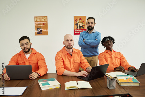 Three young adult men sitting at table using laptops with notebooks open, middle aged Caucasian man standing behind them with arms crossed, all facing camera