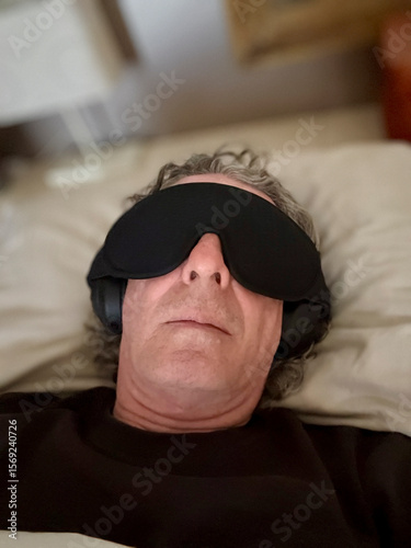 An older man lies in bed wearing a face mask and headphones, shielding himself from noise and light. Exhausted and overstimulated, he seeks refuge from the relentless symptoms of long COVID.
