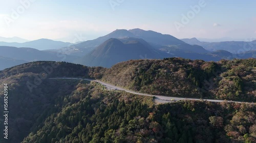 Aerial forward-moving drone shot over a winding mountain road with cars, surrounded by lush green and autumn-colored trees under a clear blue sky during the day in Japan