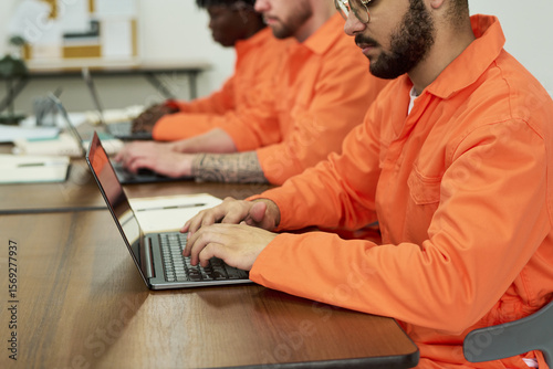 Group of young adult men of diverse ethnicities wearing orange uniforms sitting at desks using laptops, participating in educational program inside prison classroom