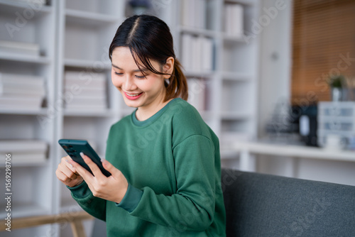 Happy Asian female student sitting on a sofa in a cozy living room, using her smartphone to browse social media while surrounded by a bookshelf filled with books Free Download Happy Asian female student sitting on a sofa in a cozy living room, using her smartphone to browse social media while surrounded by a bookshelf filled with books - GFXLoad