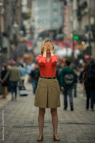 A woman stands frozen in the middle of a crowded street, covering her eyes tightly with her palms. Surrounded by the rush of people, she seems overwhelmed, trapped in a sudden panic.