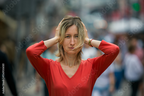 A close-up portrait of a woman standing in the middle of a crowded street, covering her ears with her hands. Her gesture creates a quiet, personal space amid the surrounding noise and movement.