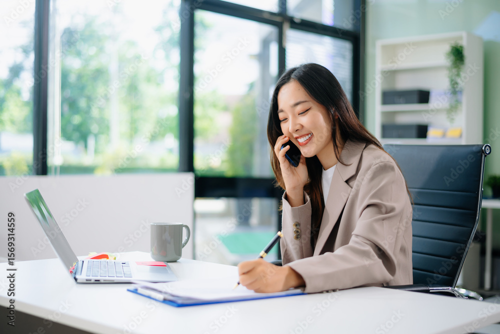 Confident young Asian businesswoman smiles while multitasking talking on her smartphone and using her laptop at a modern office desk Confident young Asian businesswoman smiles while multitasking talking on her smartphone and using her laptop at a modern office desk