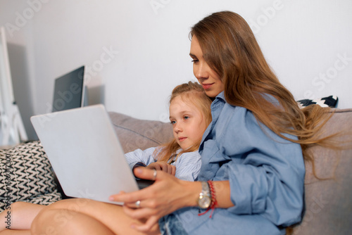 Young mother and girl watching video on laptop while sitting on sofa