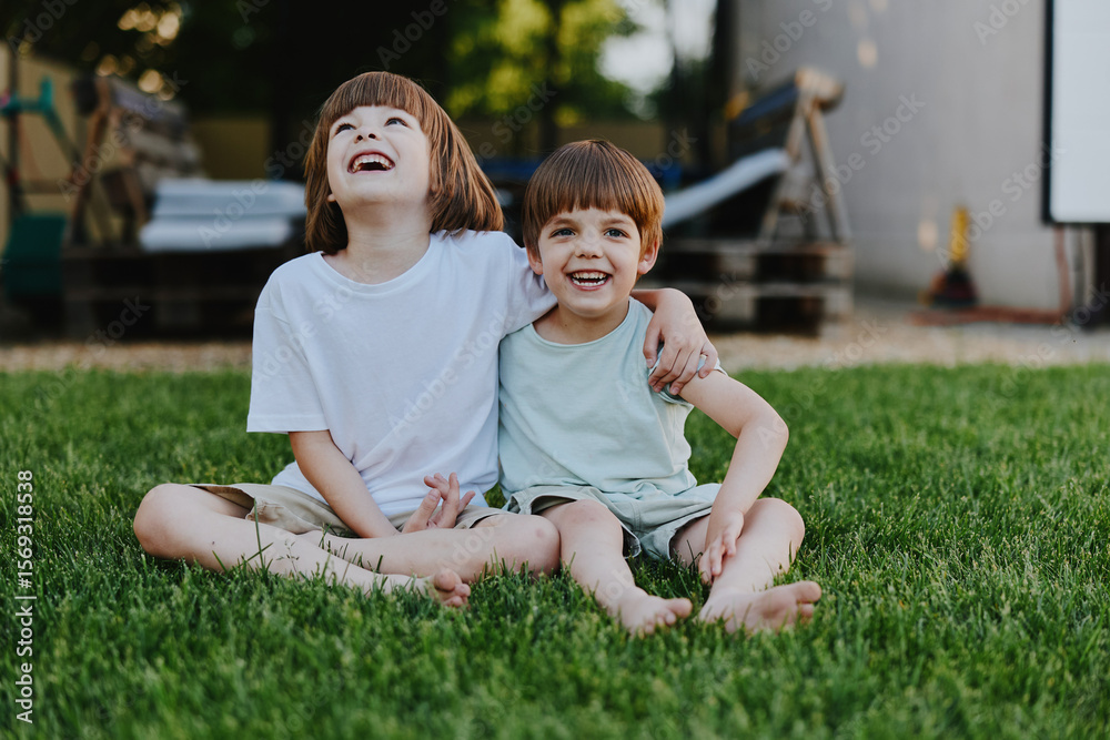 Fototapeta premium Two happy boys sitting on green grass, enjoying a sunny day while sharing laughter and friendship. A perfect moment of childhood joy captured in a natural setting.