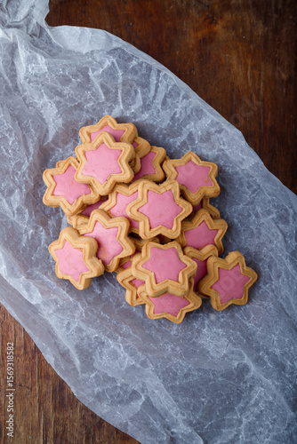 Star shaped cookies with pink frosting on white paper, wooden rustic background. Strawberry icing on cookies in shape of stars. Top view, copy space