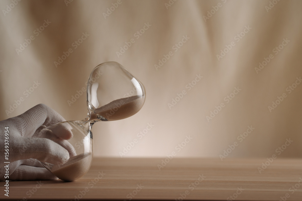 Ταπετσαρία A glove-covered hand holding a tipped hourglass on a wooden surface, symbolizing the flow of time, creativity, and patience in a contemporary minimalist style
