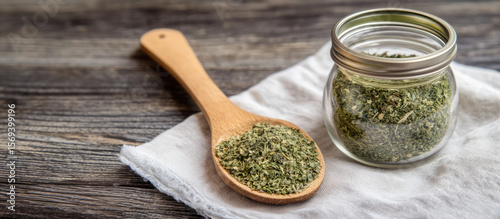 A wooden spoon with dried herbs sits on a cloth beside a small glass jar filled with the same herbs. Herbal cooking ingredient