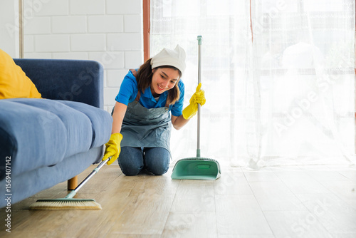 Smiling female cleaner kneels on the floor while sweeping dust and debris from under a blue sofa into a dustpan, showcasing a thorough home cleaning service in a bright, tidy living space.