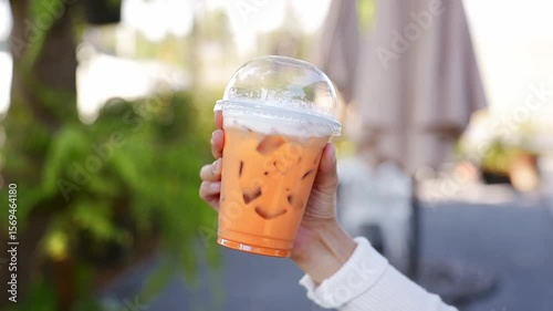 Refreshing iced drink in clear cup, vibrant orange beverage held by hand, perfect for summer outdoor enjoyment with greenery