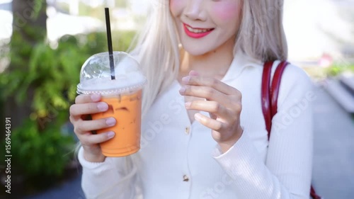 Cheerful young woman enjoys refreshing iced drink outdoors, showcasing stylish white top and long hair while smiling brightly