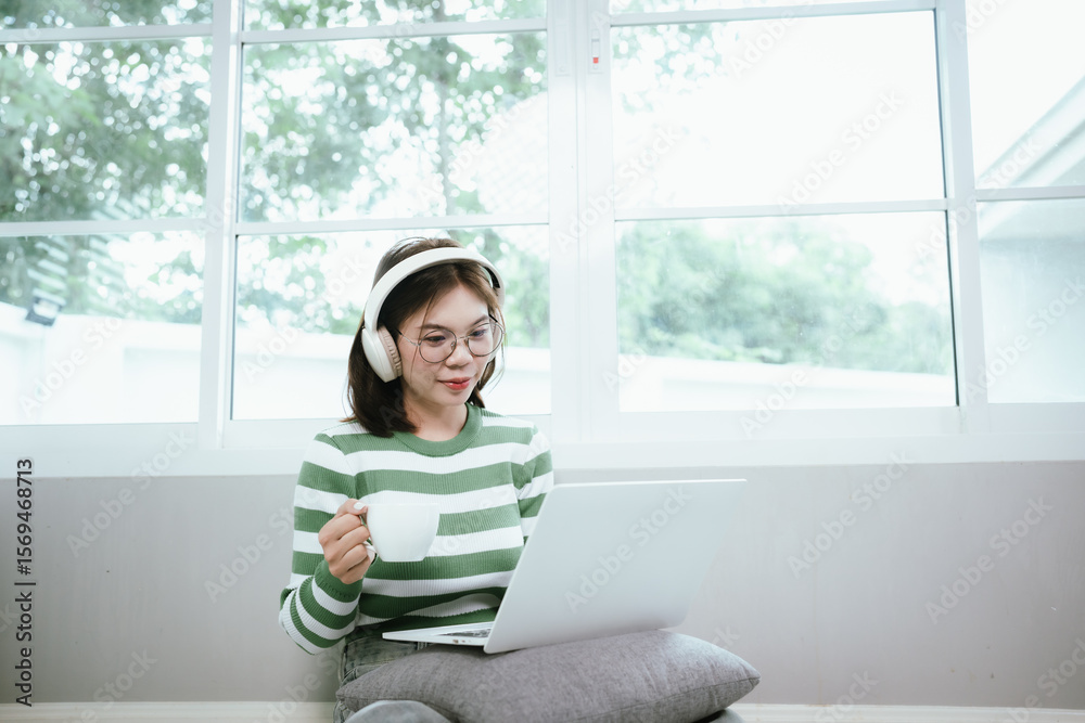 Young Asian woman lying on the carpet, wearing headphones and watching content on her laptop, enjoying a peaceful, comfortable and fun morning at home. Young Asian woman lying on the carpet, wearing headphones and watching content on her laptop, enjoying a peaceful, comfortable and fun morning at home.
