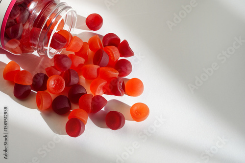 A transparent bottle lies on its side as colorful red and orange gummy vitamins spill onto a clean white surface