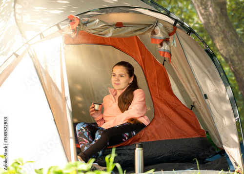 Young female traveler holding a stainless mug in a tent, enjoying the calm morning atmosphere in the forest during a solo camping trip.