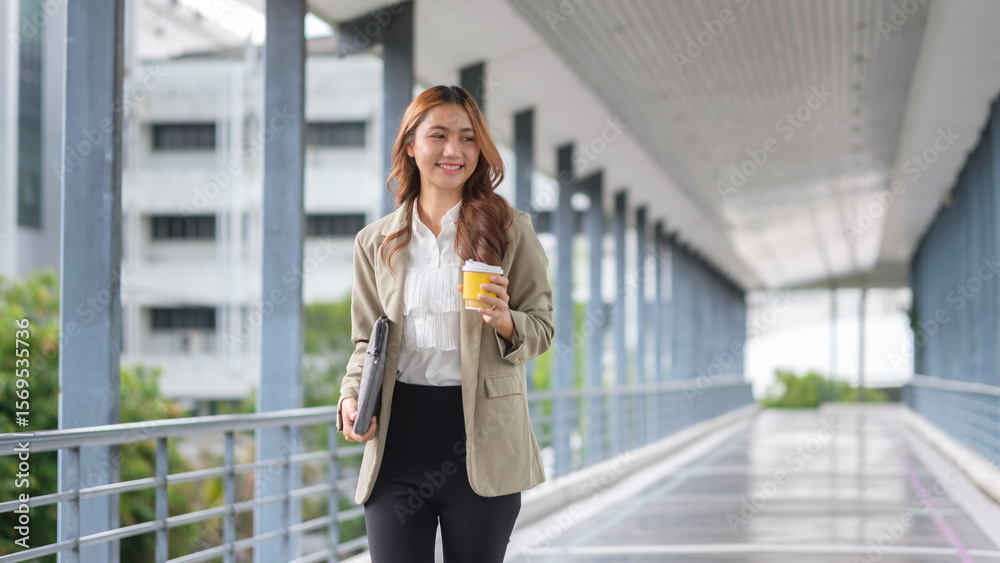 Fototapeta premium Confident young businesswoman walking across an office walkway with a coffee cup and a laptop in hand.