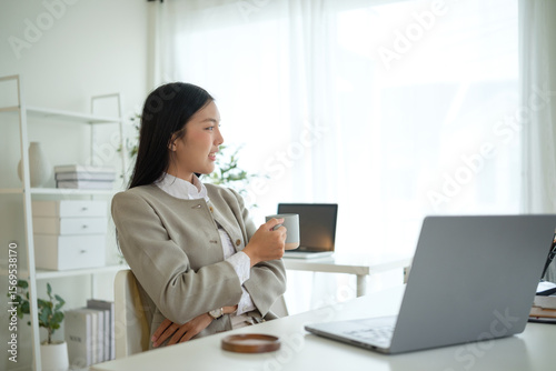Young businesswoman sitting at her desk with a cup of coffee, looking out the window in a bright modern office. Relaxing during a work break.