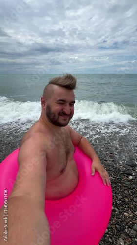 Vertical 4K travel video. Young adult white fat man with beard, stubble, pink swimming ring walks on pebble beach on Black Sea coast, takes selfie, communicates via video call. Blue sky, ocean wave