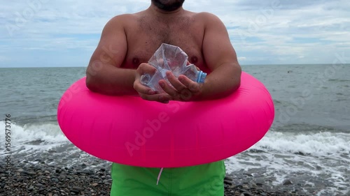 Horizontal 4K video close up. Fat white man with big belly, green shorts, pink inflatable swim ring crushes, squeezes and breaks water bottle on pebble beach. Sea. Tourism, zero waste, earth day, eco