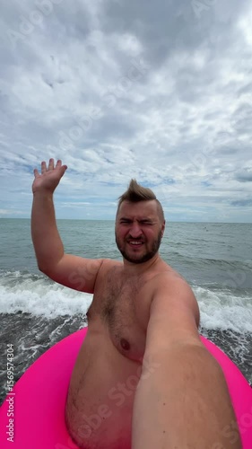 Vertical 4K video. Young adult white fat man with beard, pink inflatable swim ring waves hand, greets, talks, speaks, communicates via video call, takes selfie on pebble beach on Black Sea coast. Rest