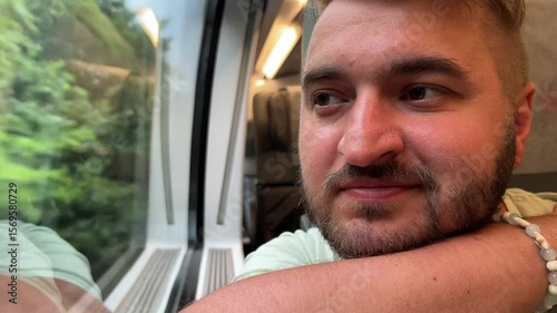 Horizontal 4K travel video close up. Young adult fat white man with stubble, green T-shirt, ring on finger sits on seat and looks out of large train window. Concept of tourism, road trip, rest, relax