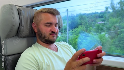 Horizontal 4K travel video. Young adult fat white man with stubble, beard, green T-shirt sits on seat and playing mobile game watching video on red smartphone. Train window. Tourism, road trip, rest