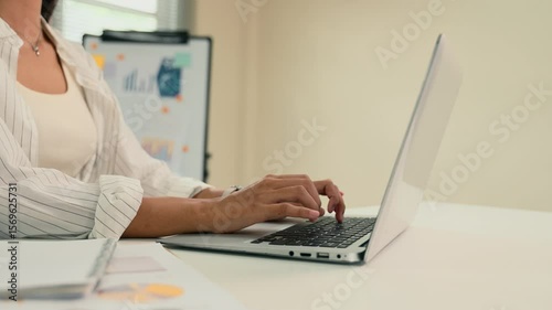 Businesswoman typing on a laptop at a modern white desk with charts and graphs in the background