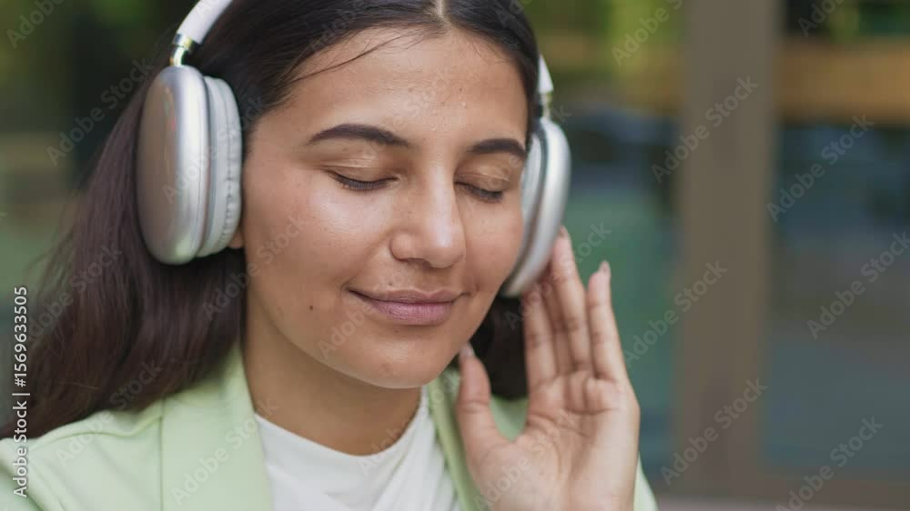 Charming woman with dark straight hair wearing headphones enjoying music with eyes closed. Slightly smiling. Feeling relaxed or immersed in sound. Touching earcup while listening outdoors. Charming woman with dark straight hair wearing headphones enjoying music with eyes closed. Slightly smiling. Feeling relaxed or immersed in sound. Touching earcup while listening outdoors.