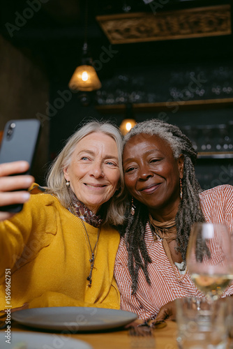 Two happy and smiling senior friends taking selfie at a table in a restaurant during a meeting, one of them is blonde, the other is African American with dreadlocks. Senior women