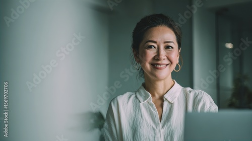 A smiling woman with dark hair pulled back, wearing a white blouse and gold hoop earrings, sits in a softly lit indoor space, exuding warmth and confidence.