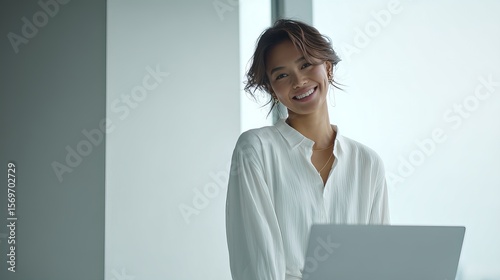 A smiling woman in a white blouse stands with a laptop, exuding confidence and professionalism in a bright, modern office setting.