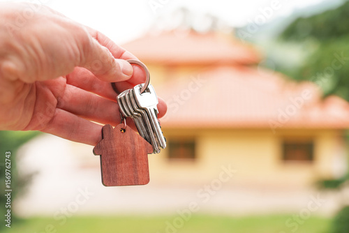 Home keys with wooden house shaped keychain against background of new bungalow in natural ecological area