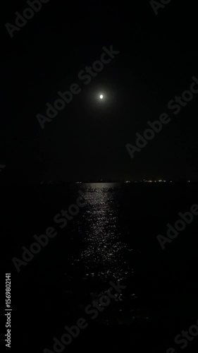 Full moon over dark sea nightscape with moonlight reflection. Peaceful ocean waves under moonlight. Tranquil night horizon with shimmering water and calm coastal lights.