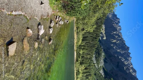 A picturesque view of a crystal-clear mountain lake with a vibrant green hue, nestled amidst a lush forest and towering rocky peaks under a clear blue sky. 