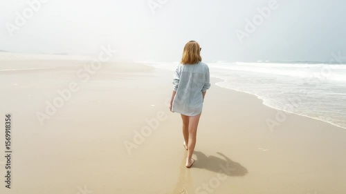 A woman walks barefoot along a peaceful beach in the misty morning light. Her relaxed pace and the soft waves create a calm, dreamy, and meditative coastal atmosphere