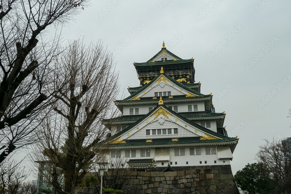 Fototapeta premium The majestic Osaka Castle stands proudly on its massive stone foundation under an overcast sky, framed by a bare winter tree, highlighting the enduring presence of this Japanese historical landmark.