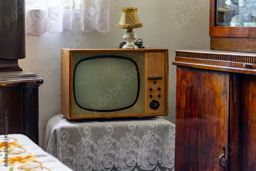 Tableau sur toile Vintage CRT TV from the communist era on a lace doily, next to a retro lamp and mid-century wooden furniture – classic 1960s–70s interior