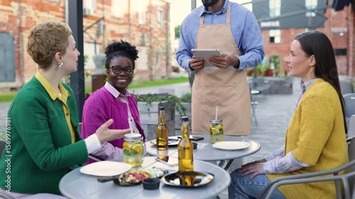 A diverse group of people enjoying a meal outdoors. A waiter takes an order on a tablet.