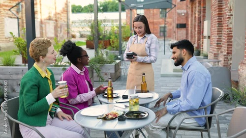 A female waiter in an apron uses a portable terminal to process a payment at an outdoor restaurant, customers are ready to pay.