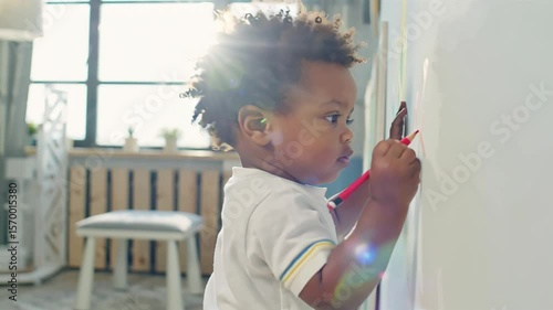 little stubborn african american boy draws with markers on the wall in the room
