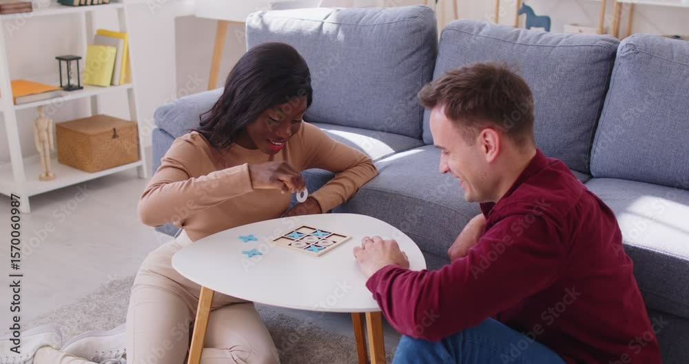 Happy interracial young couple sitting on floor in living room, playing tic-tac-toe board game together at home. Smiling man and woman enjoying and spending fun time, competing during leisure activity