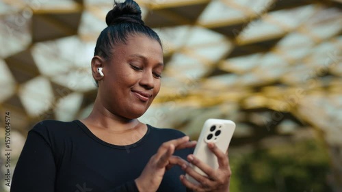 Woman Using Her Phone, Standing Outside, and Scrolling Through Social Media with Earphones In. Positive Girl Runner Scrolling Through Photos After a Run. People and Technology Concept