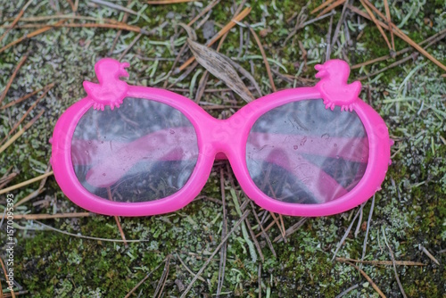 Fototapeta Naklejka Na Ścianę i Meble -  one small orange dirty plastic toy glasses lies on the ground and green moss on a summer street