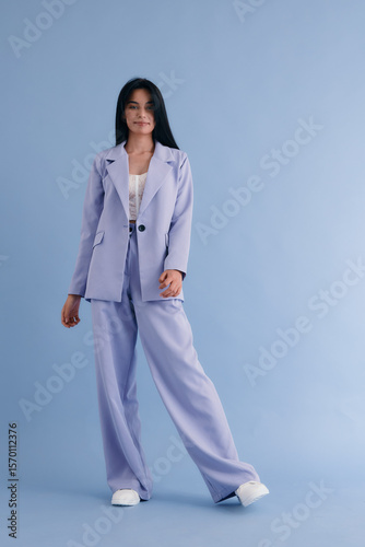 young woman in lilac pantsuit posing confidently on blue background, wearing casual white shoes and delicate lace top, standing full-length with relaxed expression