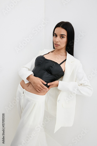 Young woman posing in a stylish black corset and white suit, standing against a plain white background with a confident and poised expression.