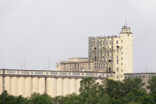 Foto Large abandoned grain silo facility with weathered towers, numerous metal pipes, and industrial structures, partially obscured by trees under cloudy skies