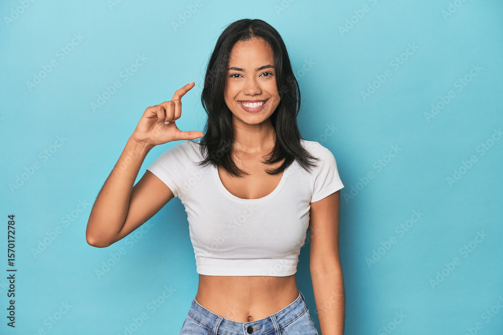 Filipina young woman on blue studio holding something little with forefingers, smiling and confident. Filipina young woman on blue studio holding something little with forefingers, smiling and confident.