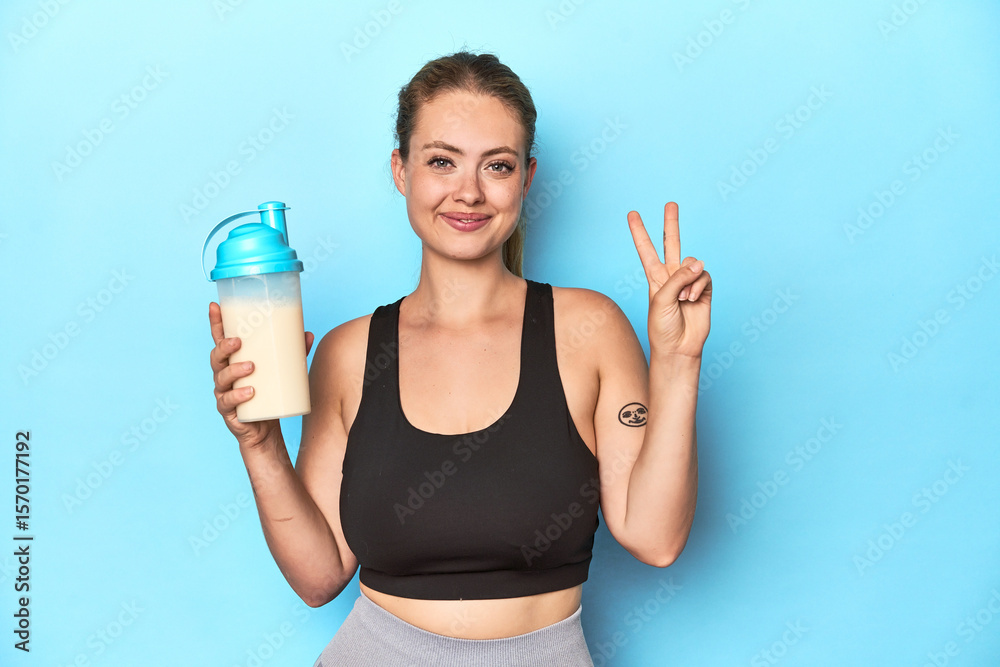 Sporty blonde with a protein shake in a studio showing number two with fingers. Sporty blonde with a protein shake in a studio showing number two with fingers.