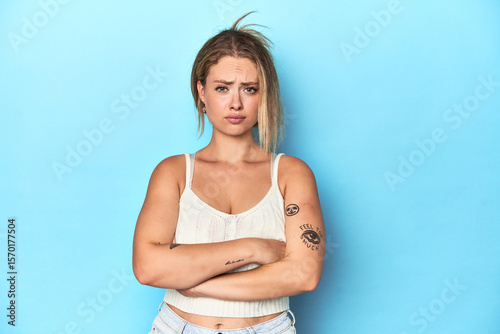 Blonde young woman in white top on blue background frowning face in displeasure, keeps arms folded.