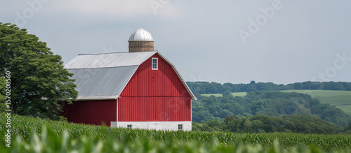 Red barn with a silver roof stands amidst lush green fields and trees in a rural setting. Scenic countryside landscape, peaceful and serene